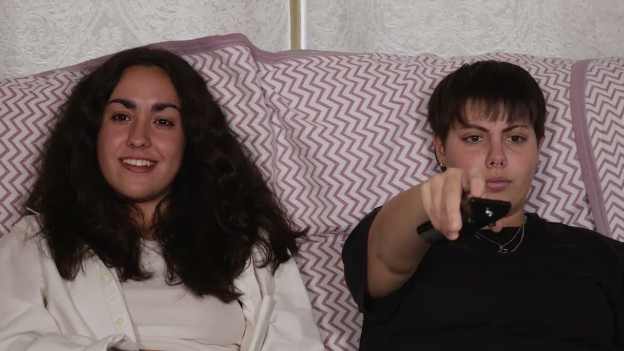 Dos chicas sentadas en un sofá; una apunta con el mando a distancia hacia la cámara