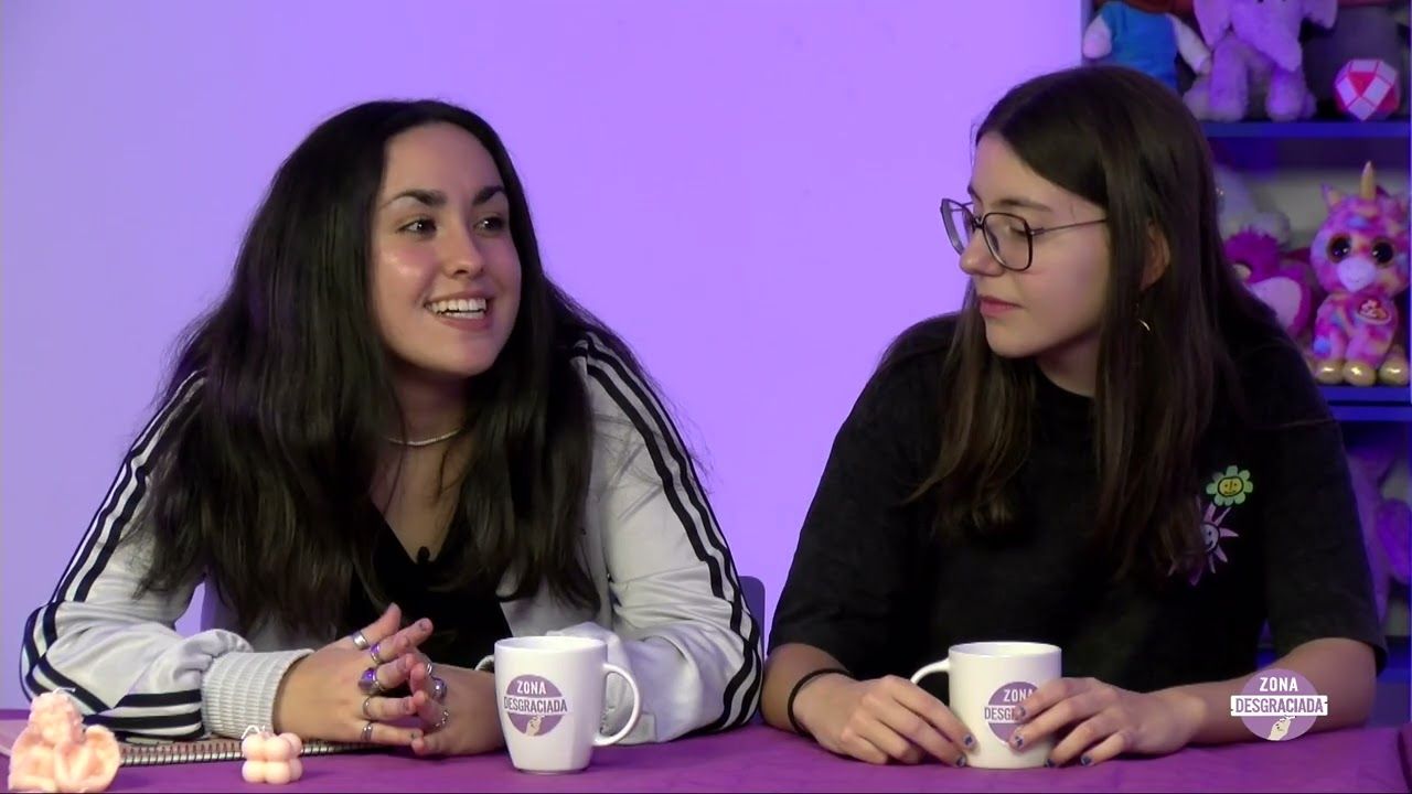 Dos presentadoras sentadas a una mesa con tazas del programa Zona Desgraciada en un plató iluminado en morado